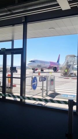 A Wizz Air plane is seen through a window at an airport. The plane is white with pink accents, and the airline's name is written on the side. A set of stairs is attached to the plane, and a person is visible on the stairs. There are signs and other equipment on the tarmac. The sky is clear and blue. The view is from inside an airport terminal, with a door and windows visible.