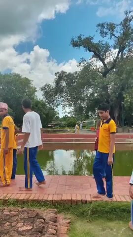 A group of people, mostly young males wearing yellow shirts and blue pants, are walking along a red brick pathway next to a body of water. Some are wearing hats. The weather appears to be partly cloudy. The area is surrounded by lush green trees and some ancient-looking stone structures. The people seem to be on a tour or an outing.