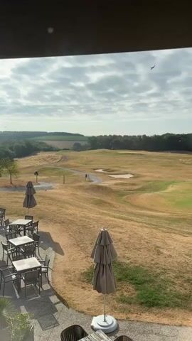 The video shows a view from a patio overlooking a golf course. There are several tables and chairs on the patio, along with closed umbrellas. The golf course is dry and brown in some areas, with green patches and a path winding through it. There is a golf cart with two people near a tee box. In the distance, there are trees and rolling hills under a cloudy sky. A bird flies across the sky briefly.