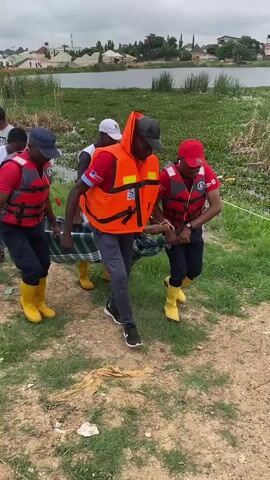 Four individuals, three wearing red vests with white markings and yellow boots, and one wearing an orange life vest over a red shirt, are carrying a person on a stretcher. The person on the stretcher is partially visible, wearing a blue and red striped garment. Two individuals in the foreground are wearing white vests with red crosses and the text "NIGERIAN RED CROSS." The environment appears to be a grassy area near a body of water with vegetation. The sky is overcast. The individuals are walking away from the water.