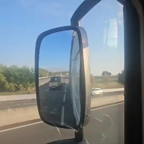 A first-person perspective video shot from a vehicle on a highway shows the view through the side mirror and the forward-facing road. The side mirror reflects a highway with traffic, including a yellow van and other vehicles, and distant mountains. The road ahead is clear with cars and trucks driving. Trees and hills line the sides of the highway, and the sky is a clear blue. In the distance, there appears to be some smoke or fog.