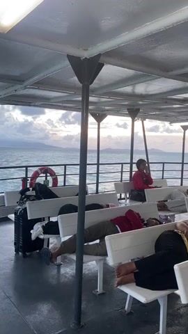 The video shows a boat interior with rows of white benches. Several people are lying down on the benches, seemingly asleep or resting. One person sits upright, appearing to be using a mobile phone. A black suitcase is visible behind the benches on the left side. The ocean and horizon can be seen through the side of the boat, suggesting it's in motion. The sky is cloudy, and the lighting indicates either early morning or late afternoon. The boat structure includes a white ceiling supported by dark gray or black pillars. A life preserver is mounted on the side of the boat.