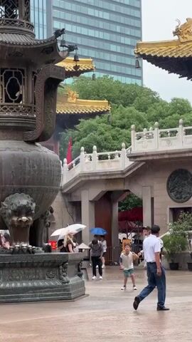 A rainy day at a temple complex. A large bronze incense burner stands in the foreground to the left. In the background, traditional temple architecture with golden roofs is visible, contrasted by a modern glass skyscraper. Several people are present. Some are holding umbrellas. A man in a white shirt and blue pants walks to the right in the foreground. A group of younger people are gathered in the middle ground, one of whom is a child. The ground is wet, indicating it is raining.