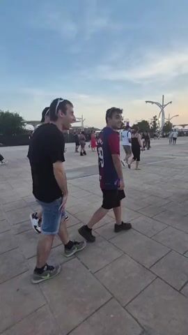 A group of people are walking across a paved area under a cloudy sky. The sky transitions from blue to a soft orange at the horizon. In the foreground, two men are walking. The man on the left wears a black t-shirt, blue denim shorts, and green and black sneakers. The man on the right wears a navy and red sports jersey with the number 69 on the back, black shorts, and black sneakers. Behind them, other people are also walking and standing, some are seen in the distance. There are structures and trees visible in the background. A large bus is parked on the right side of the frame, with people near it. The overall scene appears to be an outdoor event or gathering.