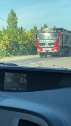 A truck is seen driving on a road. The video is taken from inside a car, looking through the windshield. The windshield is cracked. A dashboard display shows the time as 4:15, the exterior temperature as 31 degrees Celsius, and the interior temperature as 20 degrees Celsius. It also shows "Distance to Empty" as 220 km and "AUTO" for climate control. The truck appears to be a dump truck. There are trees and foliage on the side of the road, and the sky is visible.
