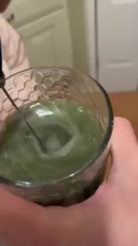 A hand holds a glass that is filled with a light green liquid, and a handheld mixer stirs the liquid. The glass has a honeycomb pattern on the sides. A person wearing a pink jacket is in the background. The scene appears to take place in a kitchen with white cabinets and wood floors.