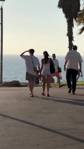 This is a vertical, medium-shot video of a couple walking hand-in-hand away from the camera, towards a body of water, likely the sea. The man is wearing a white t-shirt, khaki shorts, and a crossbody bag. The woman is in a white tube top and white shorts, carrying a black tote bag. They are on a paved walkway or road, with a grassy area and rocks separating it from the water. The sun is low in the sky, casting long shadows and suggesting it is late afternoon or early evening. The weather appears to be warm and clear, with a gentle breeze rippling the water. The sea is a calm blue, with gentle waves. In the background, other people can be seen relaxing on the grass near the water, and a palm tree stands to the right. The scene evokes a sense of leisure and a pleasant day at the beach in Tel Aviv, Israel. No visible text is present in the media.