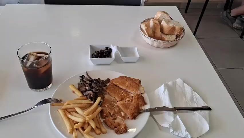 The video shows a close-up of a meal on a white table. In the foreground, a plate contains french fries, sautéed mushrooms, and a cut sandwich that appears to be breaded and filled with cheese. A fork is positioned over the fries, and a knife is resting on a white napkin to the right of the plate. To the left of the plate is a glass of dark liquid with ice, possibly cola. In the background, there are two small white dishes, one with black olives and the other empty. A basket of bread slices is to the right of the dishes. The setting appears to be a casual dining environment with a glimpse of a person's legs and shoes in the upper right corner. The camera angle is overhead and slightly angled, focusing on the food.