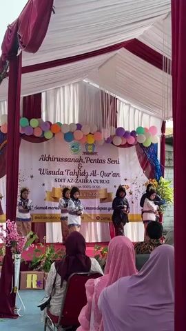 This video shows an outdoor event, likely a graduation or performance for a school, under a tent with red and white draping. The stage is decorated with balloons and a banner that reads "Akhirussanah, Pentas Seni & Wisuda Tahfidz Al-Qur'an SDIT AZ-ZAHRA CURUG Tahun Ajaran 2025/2026". There are five young girls on stage, dressed in traditional attire, some holding microphones. They appear to be performing. In the foreground, an audience is seated, mostly women wearing hijabs. The environment suggests a sunny day.