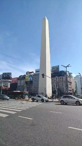 The video shows a daytime shot of the Obelisco in Buenos Aires, Argentina. Several cars pass on the road in front of the monument. Some people are visible walking around the monument. The sky is blue with some clouds.