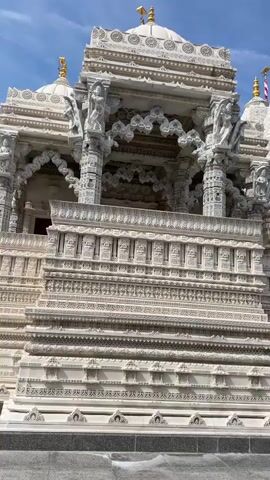 The video displays a medium shot of a white, intricately carved stone temple building. The architecture features multiple ornate towers, detailed friezes, and decorative pillars. The sky is blue with scattered clouds. The foreground shows a stone pathway and a gravel area. The camera pans slowly from left to right, showcasing the detailed facade of the temple.