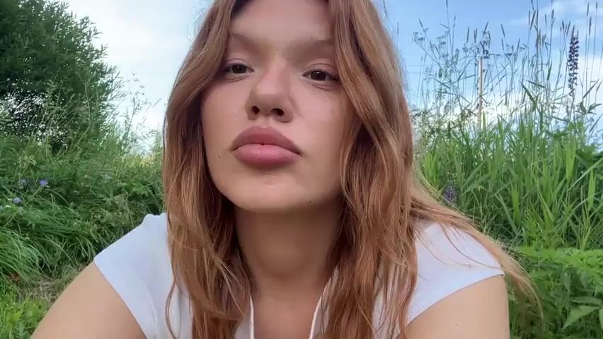 A young woman with long, reddish-brown hair and light-colored eyes is looking into the camera. She has full lips and appears to be wearing a white t-shirt. She is outdoors in a grassy area with tall reeds and trees in the background. The sky is visible and appears cloudy. In a wider shot, a person with short blonde hair, wearing an orange tank top and green shorts, is standing by a body of water, holding a fishing rod. They are surrounded by lush green vegetation.