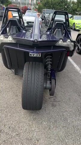 This video showcases a unique three-wheeled vehicle, a Polaris Slingshot, in a parking lot. The vehicle is dark purple with a black interior. The front of the Slingshot is prominently featured, with a large tire and suspension system visible. The camera then pans around to reveal the side of the vehicle, highlighting its sporty design and open-air cockpit. In the background, several other vehicles, including what appear to be Ford Mustangs in various colors, are parked. The scene suggests a car show or a gathering of automotive enthusiasts.

A man in a light-colored shirt and shorts is briefly visible standing near the vehicle, looking towards the camera. The overall setting is an outdoor parking lot with pavement and some greenery in the distance. The sky appears overcast, suggesting a cloudy day, possibly late afternoon or early evening given the lighting. The location is identified as Bolshoy Sochi, Russia. No specific activity or event is explicitly taking place beyond the display of the vehicle. The atmosphere seems casual and observational, focusing on the unusual vehicle. A license plate on the back of the Slingshot displays "KY 23".