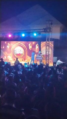 Here is a description of the video:

The video shows a man performing on a stage for a large crowd. He is wearing a blue suit and is holding a microphone. A DJ is behind him, playing music. The stage has a large screen behind it with the words "Sarkin Dariya" written on it. The audience is seated and appear to be enjoying the performance. The stage is lit up with several lights and there is a scaffolding-like structure to the right of the stage.