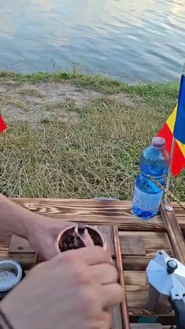 In this video, a person is grinding coffee beans outdoors by a body of water. They are using a manual coffee grinder with a wooden base and copper accents. The grinder is placed on a wooden table, next to a bottle of water and a Moka pot. Two Romanian flags are visible in the background, planted in the grass. The water is calm and reflects the sky, suggesting it is either early morning or late evening. The person's hands are visible as they operate the grinder. The scene conveys a sense of preparation for making coffee in a natural setting.