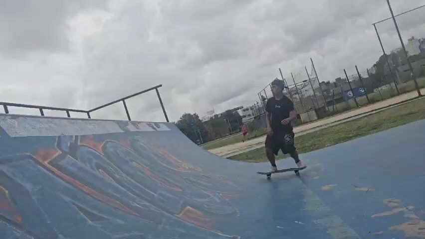 A skater wearing a black shirt, black shorts, a black cap, and white shoes is riding a skateboard on a blue skate ramp with graffiti. The ramp has a metal railing. The skater comes down the ramp and then onto flat ground. The background shows a grassy field, a chain-link fence, buildings, and a cloudy sky. A person in red walks in the background.