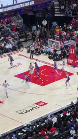 The video shows a basketball game in progress. Several basketball players are seen on the court wearing uniforms. One player in a white jersey shoots the ball and scores. A referee watches the play. Spectators are seated in the stands around the court. The court is marked with red and white lines and logos, including "TOYOTA CENTER". A digital scoreboard displays the game time as 5:40.