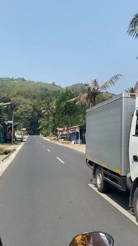 The video shows a road view from a moving vehicle, likely a motorcycle, with the rider's hand visible at the bottom of the frame. The road is paved and has white lane markings. On the right side of the road, there are small shops or stalls with red and white flags, possibly indicating national pride. One stall is painted blue. A palm tree stands tall next to one of the stalls. On the left side of the road, there is a truck parked, and further down, a pink motorcycle is visible with a person on it. The surrounding environment is lush with green trees and hills in the background, suggesting a rural or tropical setting. The sky is clear and blue, indicating a sunny day. The perspective of the video suggests a forward motion along the road, with scenery passing by.