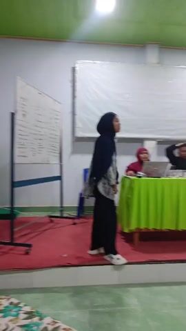 A blurry video shows a room with several people. In the foreground, a woman in a hijab walks across a red carpet. Behind her, a table is covered with a green cloth. Three people are seated behind the table, one of whom is looking at a laptop. In the background, there is a projector screen, a fan, and several windows. Further back, more people are seated at desks, some of whom appear to be students. The room has white walls and a green ceiling with lights.