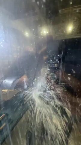 A close-up shot captures a CNC machining process. Coolant sprays vigorously from the machining area, creating a misty spray that partially obscures the view. The machine's control panel, with its display screen and numerous buttons and keys, is visible on the right side of the frame. The screen shows technical readouts and menus. A label with the text "DOOSAN PUMA" is visible on the control panel. The environment appears to be a manufacturing facility, with industrial lighting and structures visible in the background through the mist.