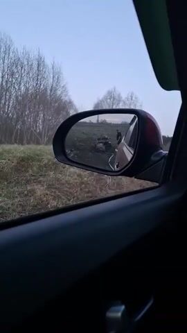 The video is shot from inside a car. In the car's side mirror, a man is visible standing next to what appears to be an overturned quad bike in a grassy field. The field is situated next to a tree line with bare trees. The sky is a pale blue, indicating it is either early morning or late evening. The car's door frame and sun visor are visible. The overall lighting is dim.