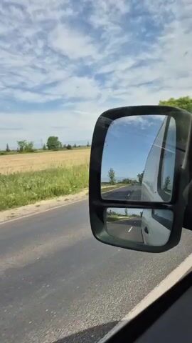 The view from inside a vehicle traveling on a road. The side mirror shows a white van behind. The road is asphalt and has yellow lines. In the background, there are green trees and a field of golden crops under a blue sky with scattered white clouds. As the vehicle moves, more cars appear in the side mirror.