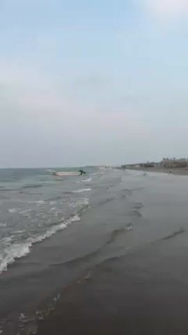 The video shows a beach with waves coming in. There is a boat in the water near the shore. In the distance, other boats and some buildings are visible. The sky is overcast. A person is on the water with some kind of motorboat.