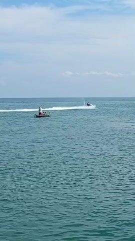 The video shows a wide shot of a body of water, likely the ocean, under a cloudy sky. In the foreground, the water is a deep turquoise. In the middle ground, a small boat with a few people on board is moving from left to right, leaving a white wake behind it. Further in the distance to the right, another jet ski is visible, also moving and creating a wake. The camera is positioned to capture the expanse of the water and the activity on it.