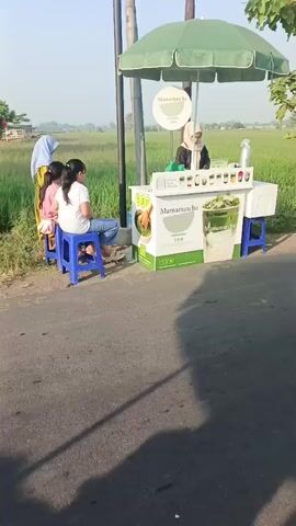 The video shows a roadside stall selling matcha drinks. Two young girls sit on blue stools at the stall, while a woman in a hijab serves a customer. A man and woman on a red motorcycle pass by the stall. In the background, a rice paddy is visible, indicating a rural setting. The stall has a green umbrella and is decorated with images of matcha drinks. The video appears to be filmed during the day.