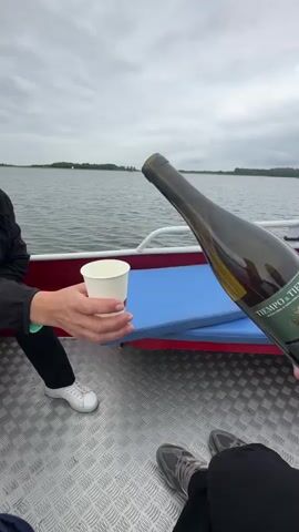 The video shows a close-up of a person's hands pouring white wine from a green bottle into a white paper cup. The cup is being held by another person's hands. The scene takes place on a boat, with a body of water visible in the background and a cloudy sky above. The interior of the boat is metallic and textured. Two people are partially visible, one on the left wearing white sneakers and the other on the right wearing athletic shoes. The label on the wine bottle reads "TIEMPO DE TIERRA".