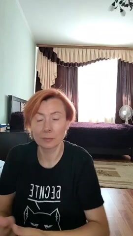 A woman with short, reddish hair is in a room, holding a hair dryer with a brush attachment. She is using the hair dryer to style her hair. The room appears to be a bedroom, with a bed, curtains, and a fan visible in the background. The woman is wearing a black t-shirt.