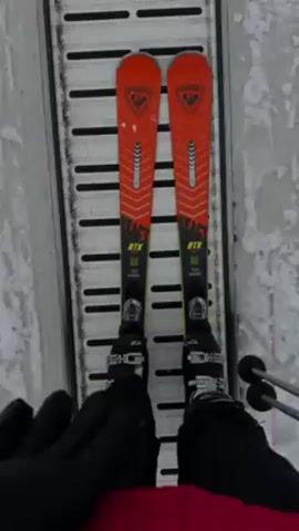 A person wearing skis is on a ski lift, going up a snowy mountain. The person is wearing black gloves, a red jacket, and orange skis that say "REACT RTX". The lift is a long metal track with holes in it. The background shows a snowy hill with some trees and other people skiing. Another person is shown ahead, also using the ski lift. They are wearing a teal jacket, pink pants, a light-blue helmet, and green skis. The weather is overcast.
