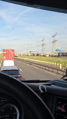 The video shows a view from inside a truck cab, looking forward through the windshield. The scene is a multi-lane highway on a clear, sunny day, likely late afternoon or early evening given the warm light. There is significant traffic congestion, with vehicles backed up for a considerable distance.

Prominently featured are several large trucks, including those carrying shipping containers. One red container is clearly visible, labeled "CAI." Other vehicles include cars and vans. The setting appears to be an industrial area, indicated by the numerous high-voltage power lines and pylons visible in the background, as well as what looks like a port or large logistics facility with more containers and cranes in the distance.

A blue road sign is visible on the left side of the highway, providing directional information. Text on the sign indicates "Strand Maasvlakte" at the top. Below this, it lists destinations and routes: "Distriparks Maasvlakte," "Oostvoome (N205)," "Rotterdam (A15)," and "Oostvoorne (N205)." The presence of "Maasvlakte Rotterdam" on the sign strongly suggests the location is Maasvlakte, a port area within Rotterdam, Netherlands.

The overall mood conveyed is one of slow progress due to traffic. The clear sky and bright sunlight suggest fair weather. No people are explicitly visible in detail, though the occupants of the other vehicles are implied. The focus is on the road, the traffic, and the industrial landscape. The truck's dashboard is partially visible at the bottom of the frame, with a speedometer showing a speed between 0 and 10 km/h, consistent with being in a traffic jam.