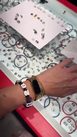 The video shows a close-up of a person's wrists adorned with multiple bracelets, followed by a panning shot of a display case filled with various charms and bracelet cords. The display case contains numerous small, colorful charms with different designs, including flags, symbols, and figures. Bracelets with braided cords in red, blue, and black are also visible, along with individual charms scattered on the white surface of the display. In the background, two individuals wearing white shirts with "STAFF" badges are partially visible. One person is holding a small golden object. The scene appears to be a retail environment, possibly a jewelry store or a market stall, where customers can create personalized bracelets.