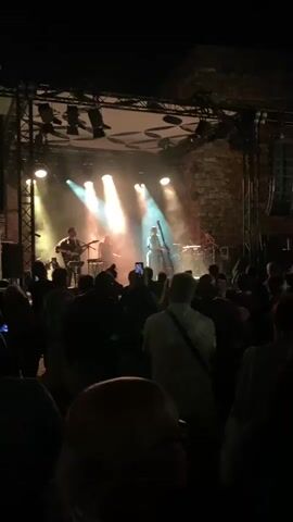 This is a dimly lit, upward-angled video of a live music performance at night. The stage is set up with multiple spotlights illuminating a band playing. From left to right, there is a guitarist standing and singing, a keyboardist seated, a double bass player standing, and a drummer seated behind a drum kit. The band is performing on a stage in front of a stone wall structure, possibly a castle or old building. In the foreground and midground, a crowd of people are gathered, facing the stage and watching the performance. Many of them are silhouetted against the stage lights. Some audience members are holding up their phones to record or take pictures of the band. The atmosphere appears to be that of an outdoor concert or festival.