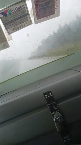 The video shows the inside of a vehicle, likely a car or truck, with a view of the windshield. Rain is falling heavily, blurring the view outside. There are several stickers on the windshield, including one for "Eastlake Insurance."  A security bar with a padlock is attached to the dashboard, preventing the steering wheel from being turned. Music is playing in the background. The vehicle appears to be stationary.