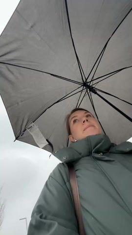 A woman stands under a large, gray umbrella. She is wearing a green puffer jacket and has a strap of a bag across her chest. The camera is positioned looking up at her. The sky is overcast. A sign with "GALERIA" is visible in the background. The woman looks up and around.