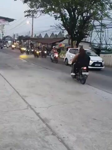 A group of motorbikes are riding down a street. The motorbikes are all going in the same direction. There are people on the motorbikes. There are also some cars and trees on the side of the road. The sky is overcast. The street is paved and there is a sidewalk on the side of the road.