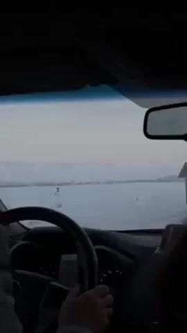 The view from inside a car shows a driver navigating a snow-covered landscape. The sky is overcast, and the distant horizon is slightly hazy. A triangular yield sign stands on the side of the road, which is entirely covered in snow. The driver's hands are visible on the steering wheel, and a passenger is partially visible in the right side of the frame. The car's dashboard is also visible.