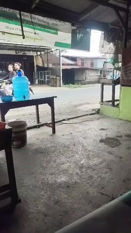 The video shows a street scene from inside a covered area, possibly a shop or home. In the foreground, there is a wooden table with a blue water jug and a blue bowl on it. Next to the table is a white bucket. The ground is concrete. A motorcycle with one rider passes by on the street in the background. A banner is visible hanging above the scene. The environment appears to be outdoors in a moderately lit area.