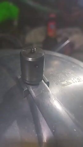 The video shows a close-up of a pressure cooker's valve. Steam is seen escaping from the valve, indicating that the pressure cooker is in use. The valve appears to be made of metal and has a threaded screw at the top. The pressure cooker is placed on a surface, and the background is blurred, showing some objects and a person. The lighting is dim, suggesting an indoor setting, possibly a kitchen.