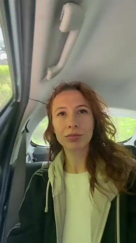 A young woman with reddish-brown hair, wearing a white shirt under a light gray hoodie and a black jacket, is sitting in the back seat of a white car. The car is moving along a road lined with green trees on a sunny day. She looks at the camera briefly, then leans out the open car window, letting the wind blow through her hair.