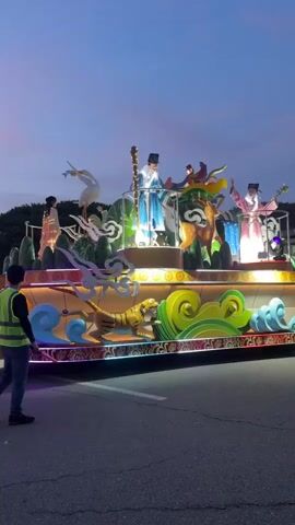 The video shows a street at dusk, possibly during a parade or festival. A large decorated float is visible on the right, featuring figures in traditional attire and illuminated elements. Several people in safety vests are directing the activity. On the left side of the street, a group of performers dressed in colorful, illuminated costumes are walking. Some are wearing mascot heads. The sky is a soft gradient of blue and pink, and streetlights and building lights are beginning to illuminate the scene. The ground is asphalt, marked with white lines. The overall atmosphere appears festive and engaging.