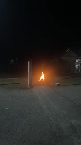 A nighttime outdoor shot captures a small fire burning in the center of a dirt path. To the left of the fire, a concrete pillar stands tall. In the background, a dimly lit structure with a sloped roof is visible. The ground is covered in dirt and small stones, and a few scattered white objects, possibly bricks or stones, are present on the ground. The sky is dark and devoid of stars, indicating nighttime. The fire casts an orange glow on its surroundings, illuminating the immediate area with a warm light. The overall scene appears to be rural or undeveloped, with a rough, unpaved road leading towards the fire. The focus is on the burning fire and the surrounding environment.