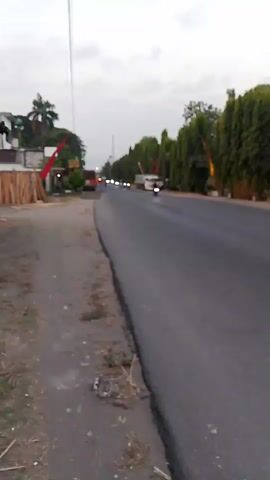 The video shows a road with a shoulder on the left side. On the shoulder, there are stacks of lumber and a building. A few flags are visible on the left side of the road. The road itself is paved and appears to be a two-lane road. There are several vehicles on the road, including motorcycles and what appears to be a truck. The road is lined with trees on the right side. The sky is overcast. The video is filmed from a low angle and is slightly blurry.