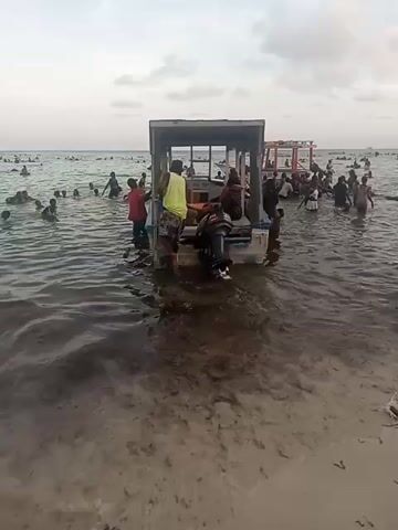 People wade in shallow water surrounding a small boat with an outboard motor near a beach crowded with other people. Several individuals are seated in the boat. One man, wearing a yellow tank top and camouflage shorts, stands on the boat's edge. Another person in dark clothing is reaching out from the boat towards someone in the water. The boat has a simple roof structure with open sides. A second, larger boat is visible further out in the water. The water is murky, and the sky is overcast.
