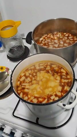 The scene shows two pots of soup cooking on a white stovetop. The pot in the foreground is white with a black rim and handles, filled with a brownish broth and small, white, diced vegetables. Steam rises from the soup. The pot in the background is stainless steel and also contains broth and the same diced vegetables. Beside the first pot is a glass jar with a yellow lid. A set of utensils sits on a plate to the left of the jar. The environment suggests a kitchen setting.