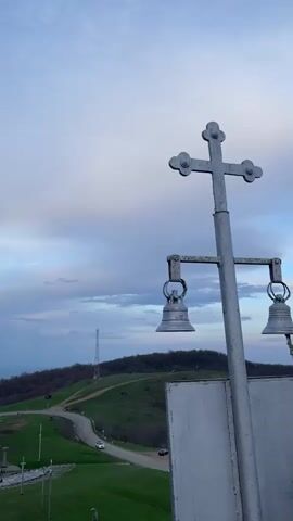 The video shows a bell being rung at a high altitude location. The view then pans around to show a concrete wall, a religious shrine, and crosses. The last shot shows a view of a green landscape with a cloudy sky behind metal religious symbols.