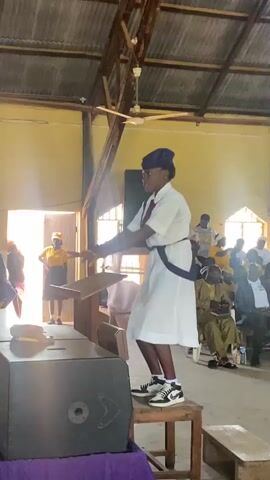 The video shows a young woman standing on a wooden chair in what appears to be a church, gesturing as if conducting an orchestra. She is wearing a white uniform dress with a navy blue sash and cap. Her sneakers are black and white. In front of her is a wooden lectern with a sheet of music on it. Behind her, people are seated in pews, some wearing traditional attire. The building has a corrugated metal roof and yellow walls. There is a large speaker near the woman and an open doorway in the background.