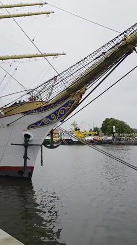 The video shows the bow of a large white sailing ship docked in a harbor. The ship has "DEUTSCHLAND" written on its side. An anchor hangs from the bow. In the background, other boats and a Ferris wheel are visible. The water is calm and the sky is overcast.